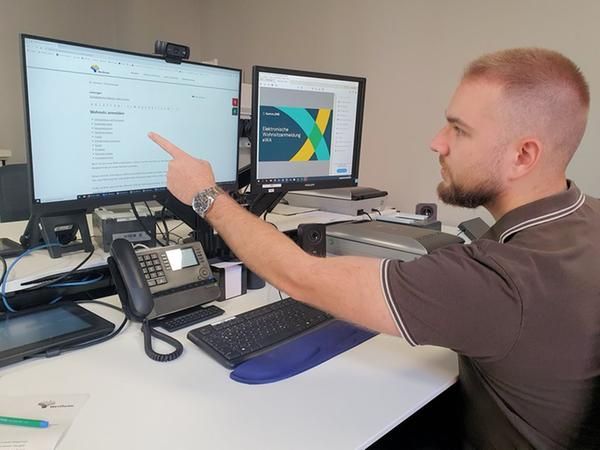 Ein Mann sitzt an einem Schreibtisch und zeigt auf einen von zwei Bildschirmen